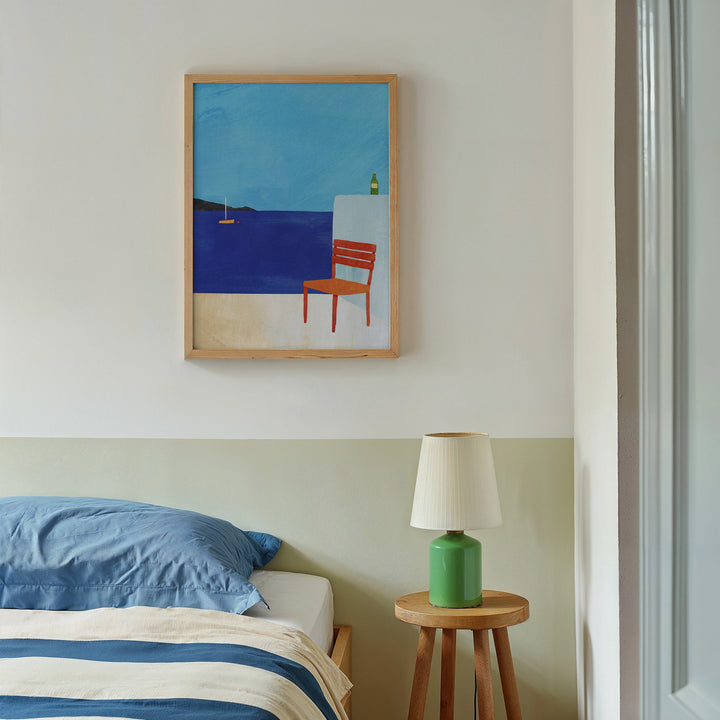 Bedroom with a painting of a chair by the sea on the wall, a green lamp on a wooden side table, and blue bedding.