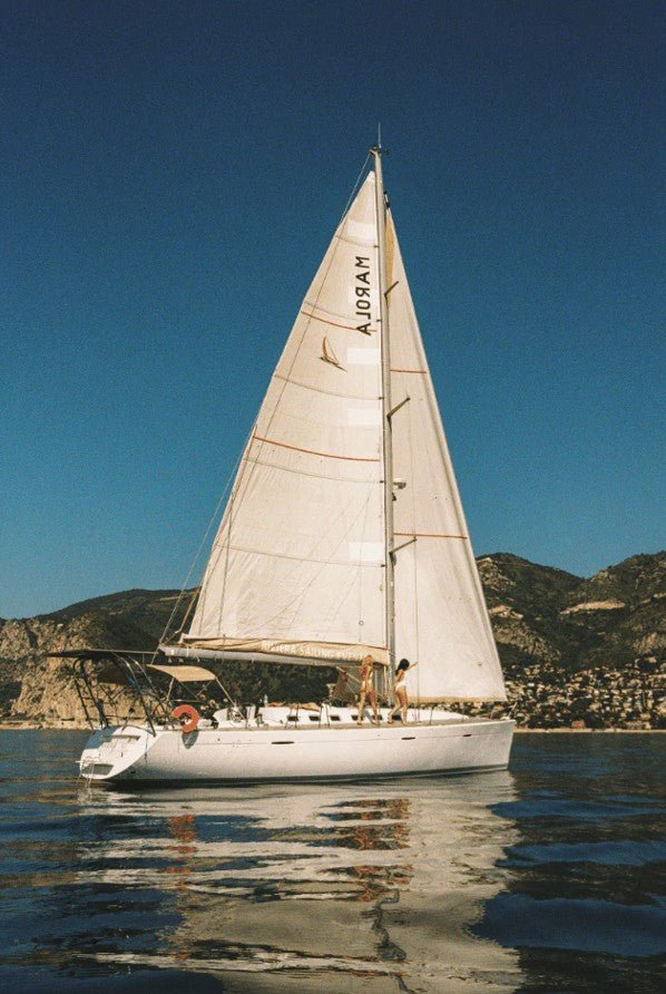 A white sailboat with a tall mainsail is shown on clear, slightly rippled water, with two figures standing on deck, against a backdrop of steep, dry coastal hills and a clear blue sky. framed