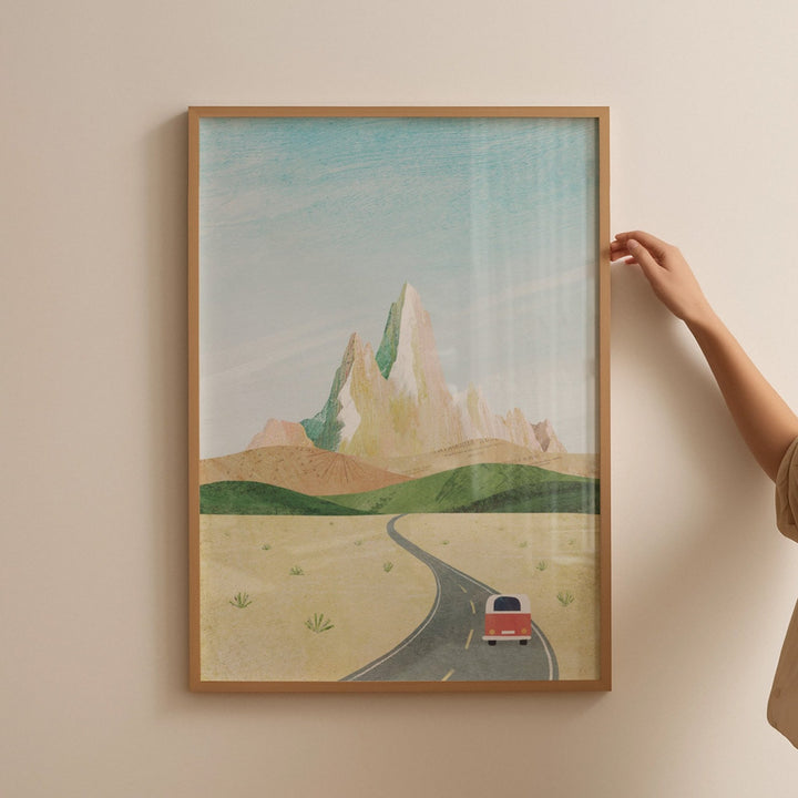Artwork of a desert scene with a road and car, held by a person against a beige wall.