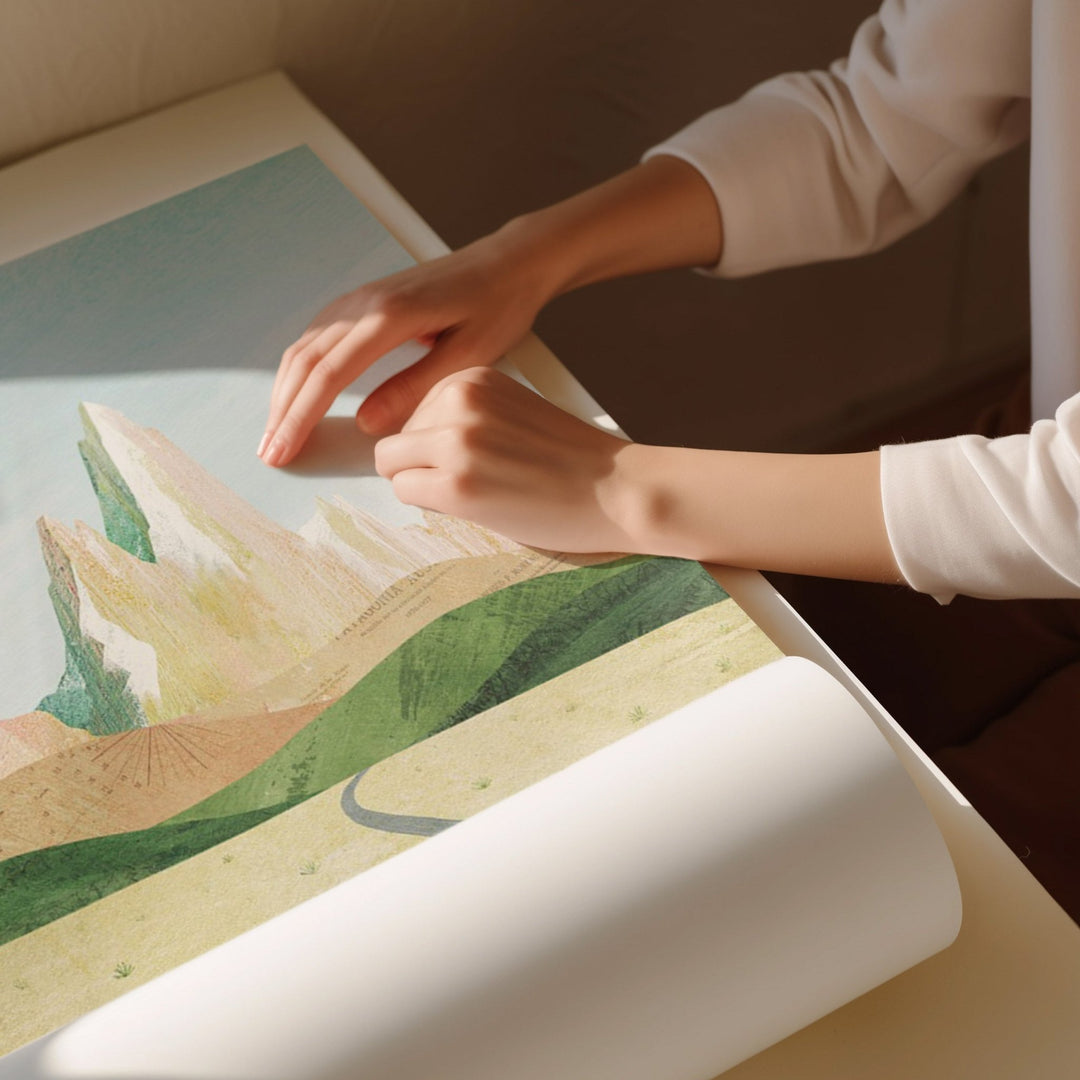 Person holding an artwork with a scenic design on a table.