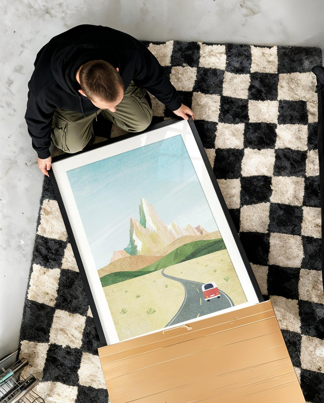 Person holding an artwork of a landscape with a car on a checkered rug.