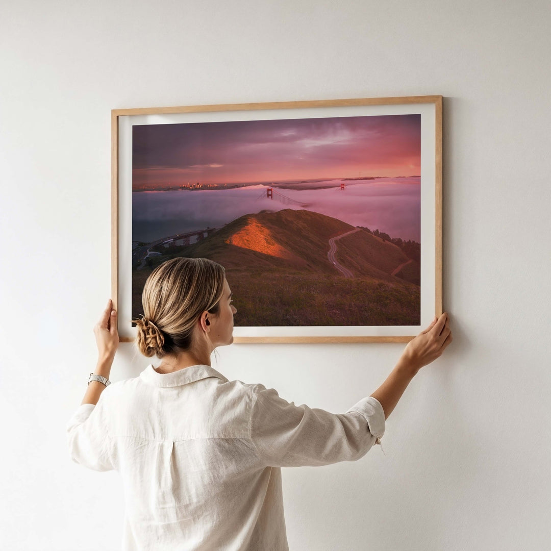 Person hanging a photograph of a sunset over a bridge on a white wall