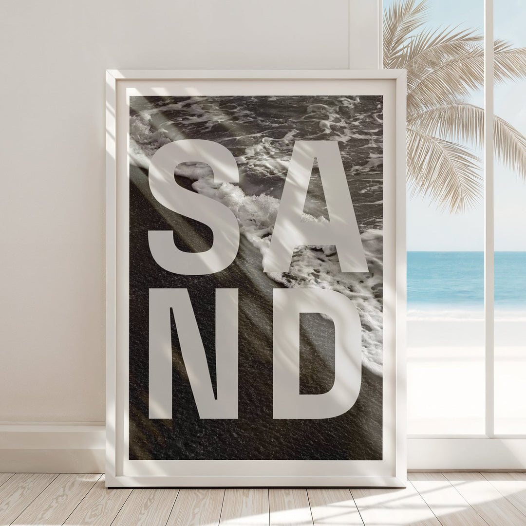 Artwork with 'SAND' text in a beach setting, displayed in a room with a window showing palm trees and ocean.
