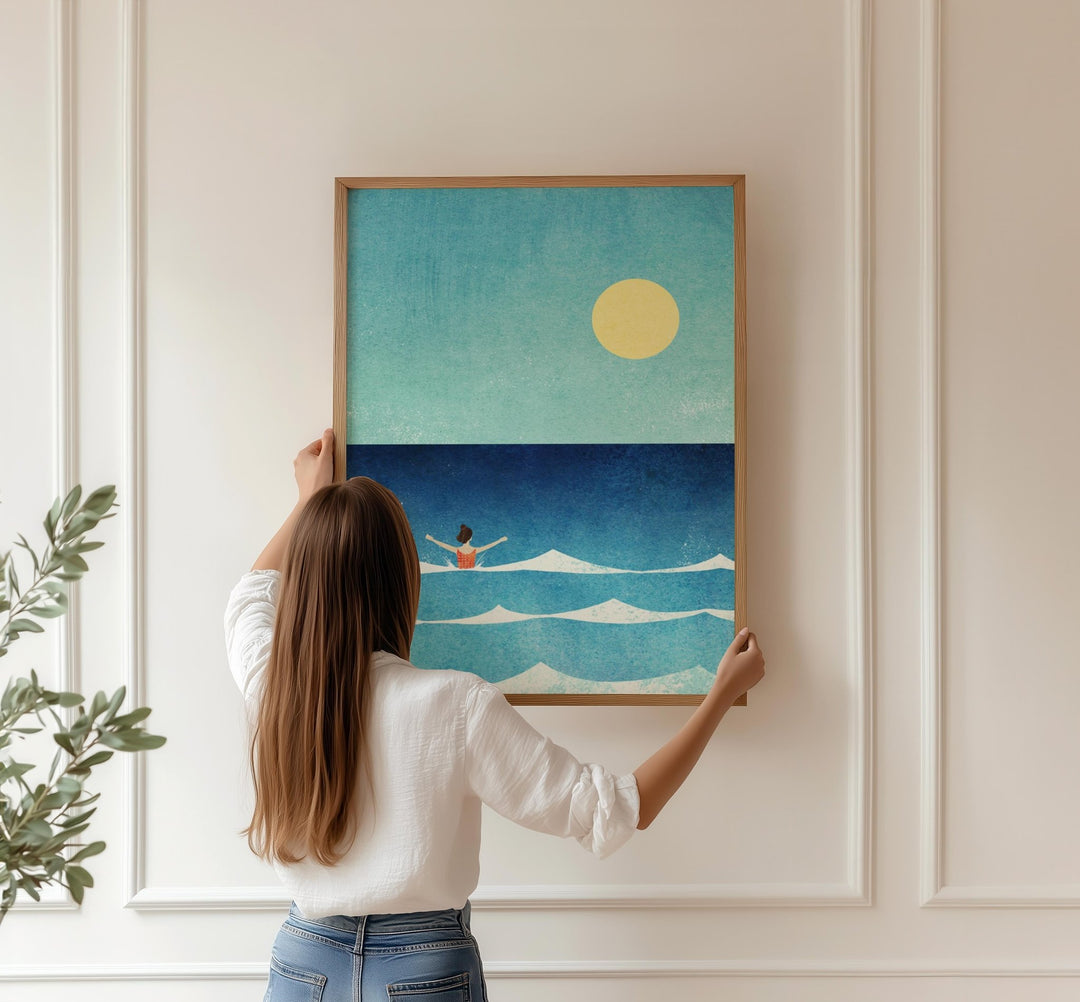 Woman hanging an artwork of a person in the ocean with a yellow sun on a white wall.