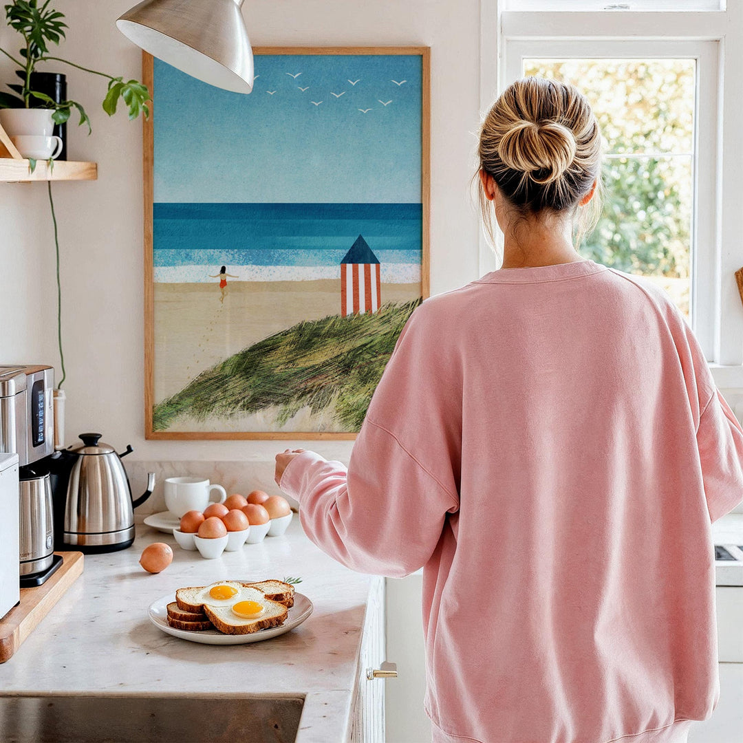 Person in a pink sweater standing in a kitchen with a beach scene painting on the wall.