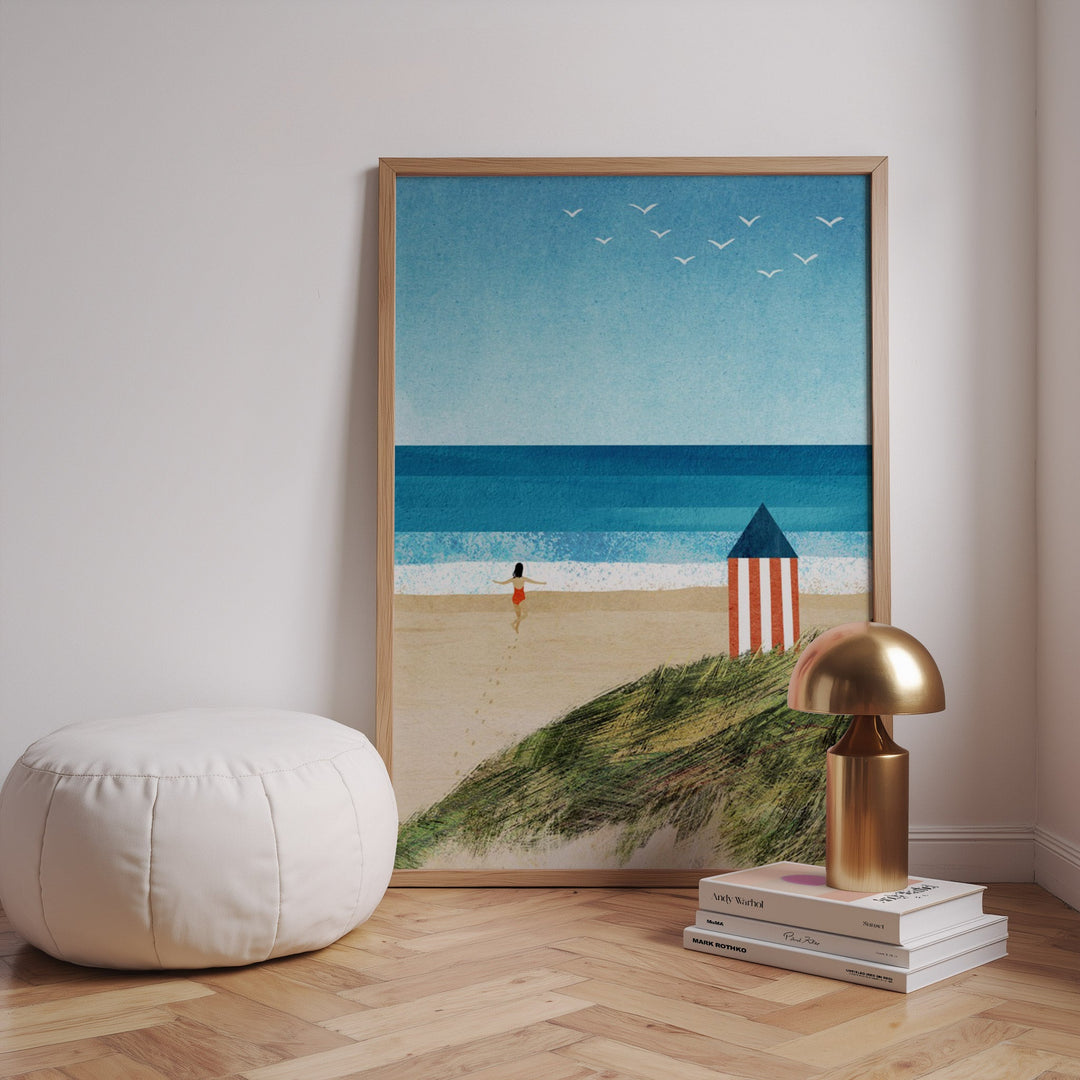 Beach scene art print on a wall with a lamp and books on a wooden floor.