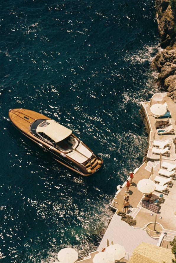 An overhead photograph captures a sleek, mahogany-trimmed wooden speed boat floating on dark blue, sun-dappled water next to a luxurious seaside dock where several white sun loungers and pale umbrellas are set up. framed