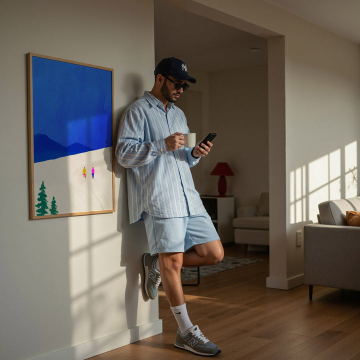Man in a living room using a smartphone with a cup, wearing a cap and light clothing.