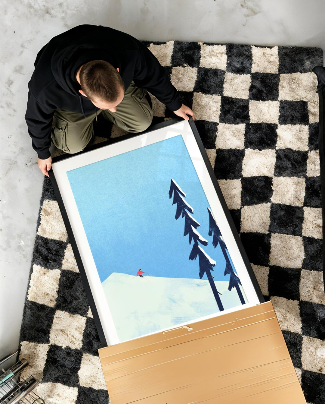 Person unboxing an artwork of trees on a mountain with a checkered rug underneath.