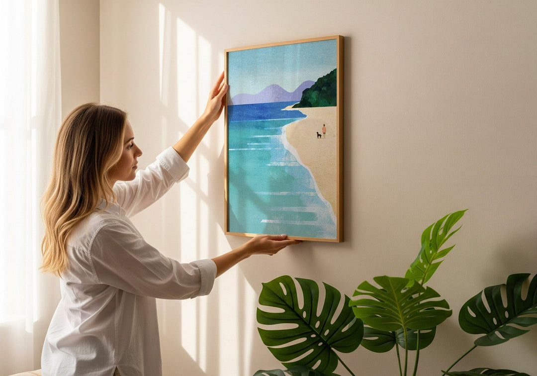 Woman adjusting a beach painting on a wall with plants in the foreground.