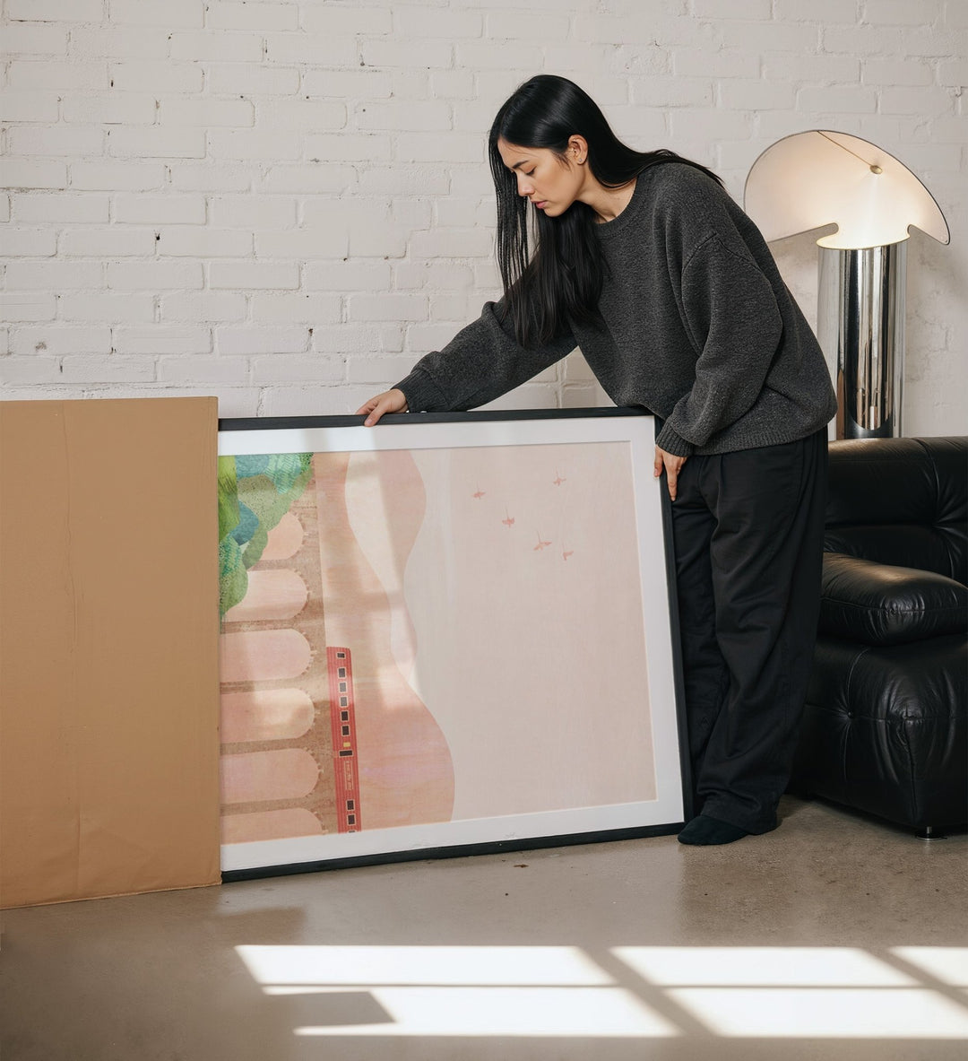 Person unpacking a picture from a cardboard box in a room with a white brick wall and a lamp.