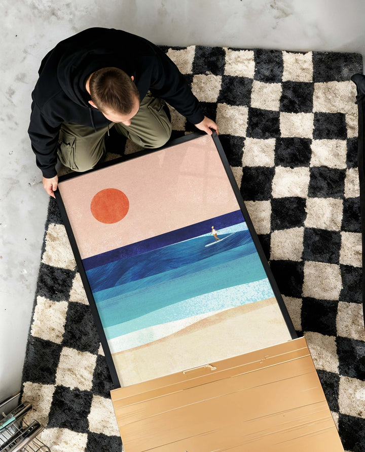 Person unboxing artwork of a beach scene with a sun and sailboat on a checkered rug.