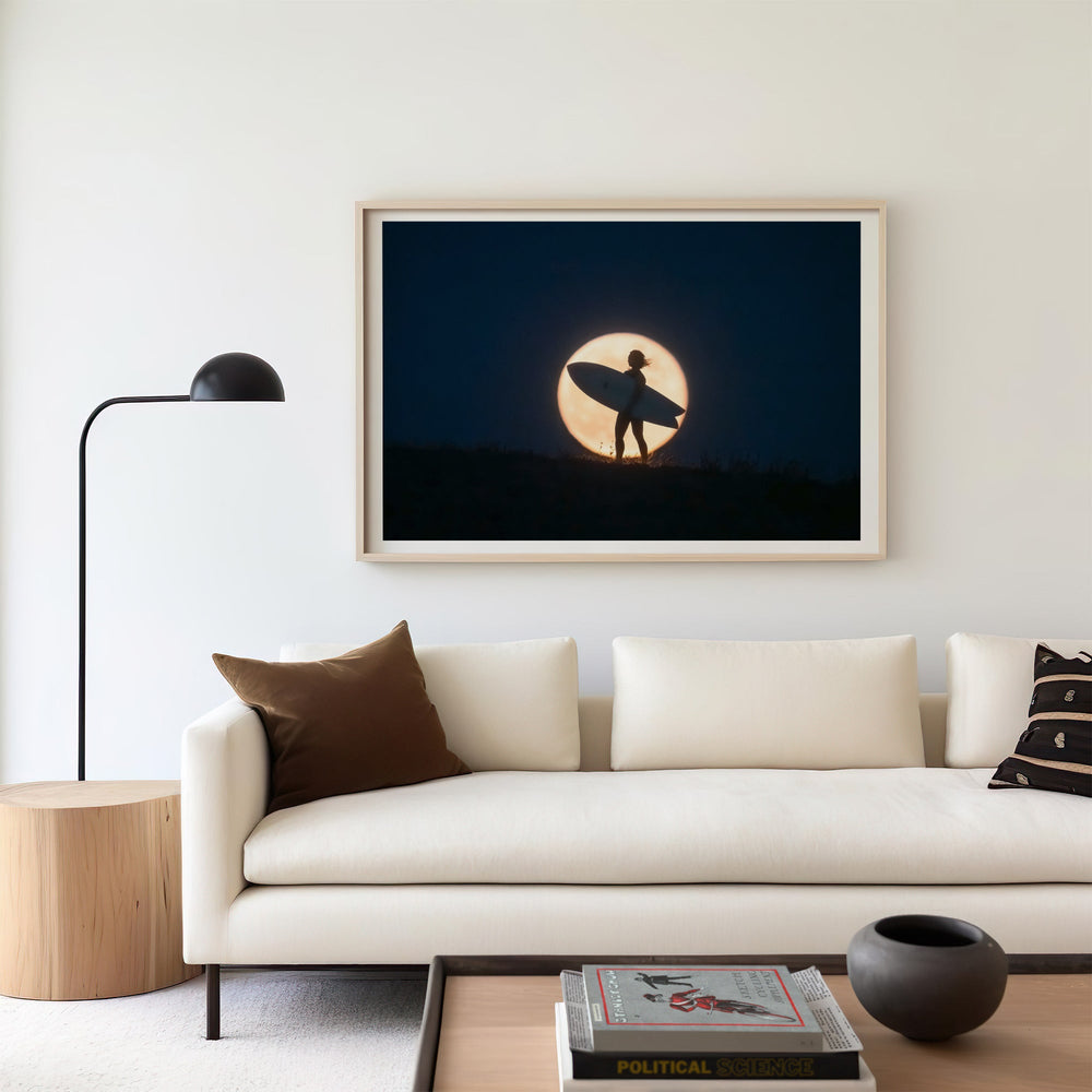 Artwork of a silhouette holding a surfboard against a moonlit sky above a sofa in a living room.