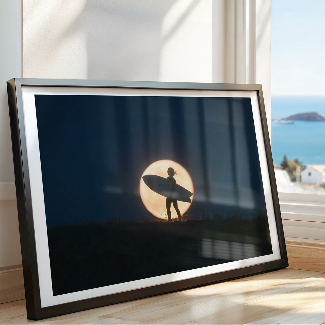 Artwork of a silhouette holding a surfboard against a sunset sky, placed on a windowsill with a view of the ocean.