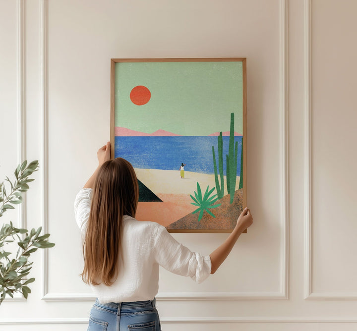 Person hanging  artwork of a desert scene with a red sun on a beige wall.