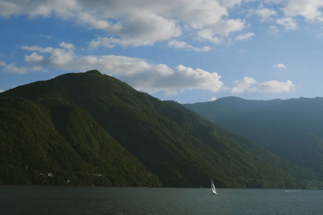 A wide landscape photograph captures a single, small white sailboat on dark water below towering, steep, forested green mountains beneath a bright blue sky with white clouds. framed