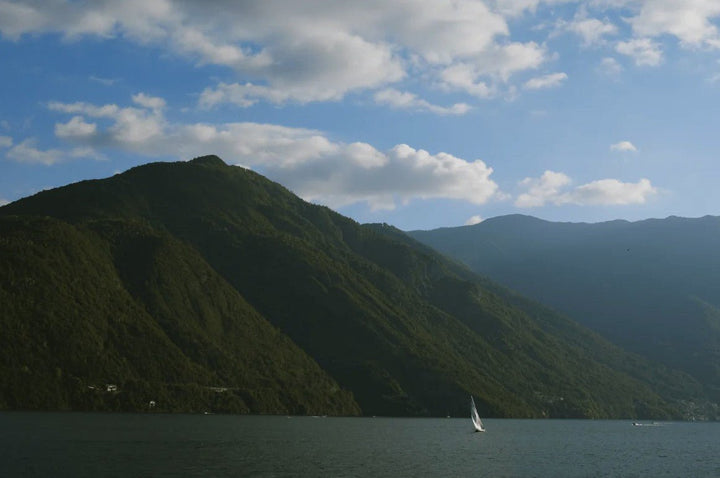 A wide landscape photograph captures a single, small white sailboat on dark water below towering, steep, forested green mountains beneath a bright blue sky with white clouds. framed