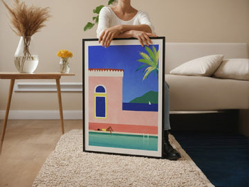 Person holding an artwork of a colorful coastal scene in a living room.