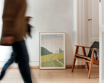 Person walking past landscape print on a wall in a room with wooden flooring.