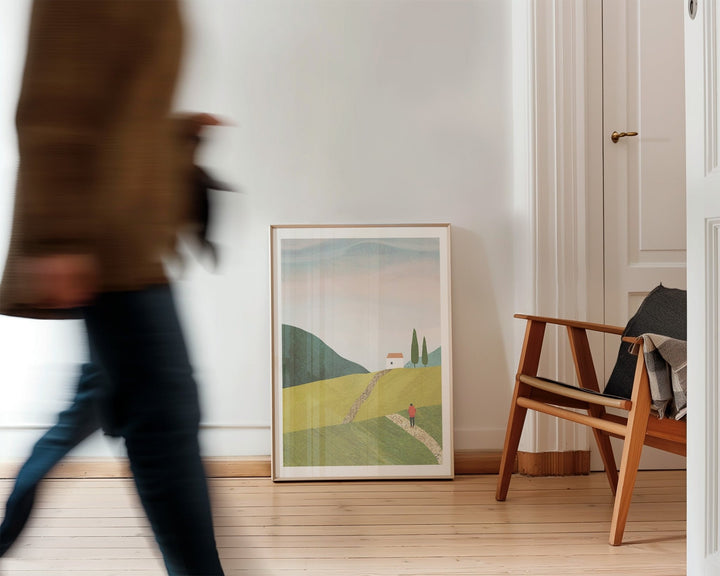 Person walking past landscape print on a wall in a room with wooden flooring.